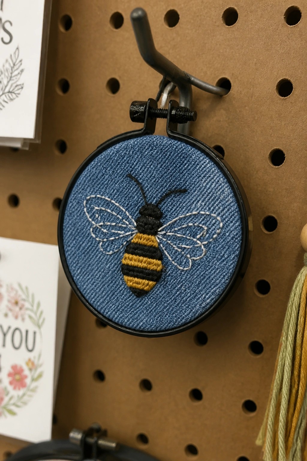 Black embroidery hoop with yellow-and-black bee stitched on blue denim, hanging on pegboard