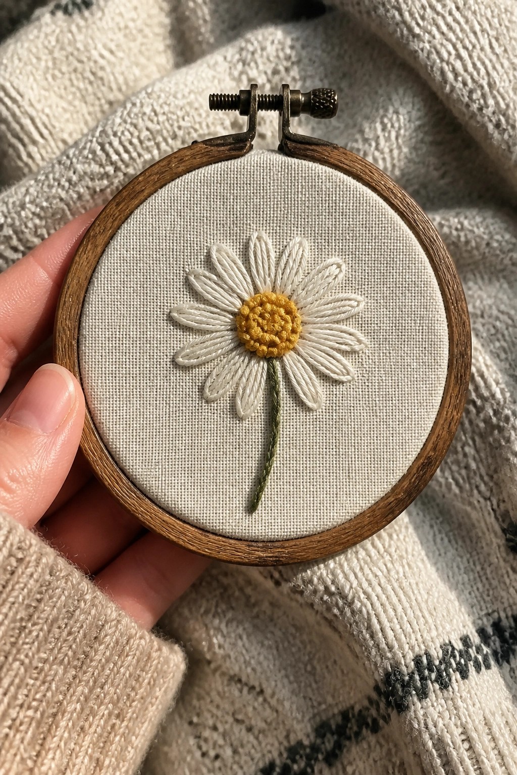 Hand holding wooden hoop with embroidered daisy of white petals and yellow center.