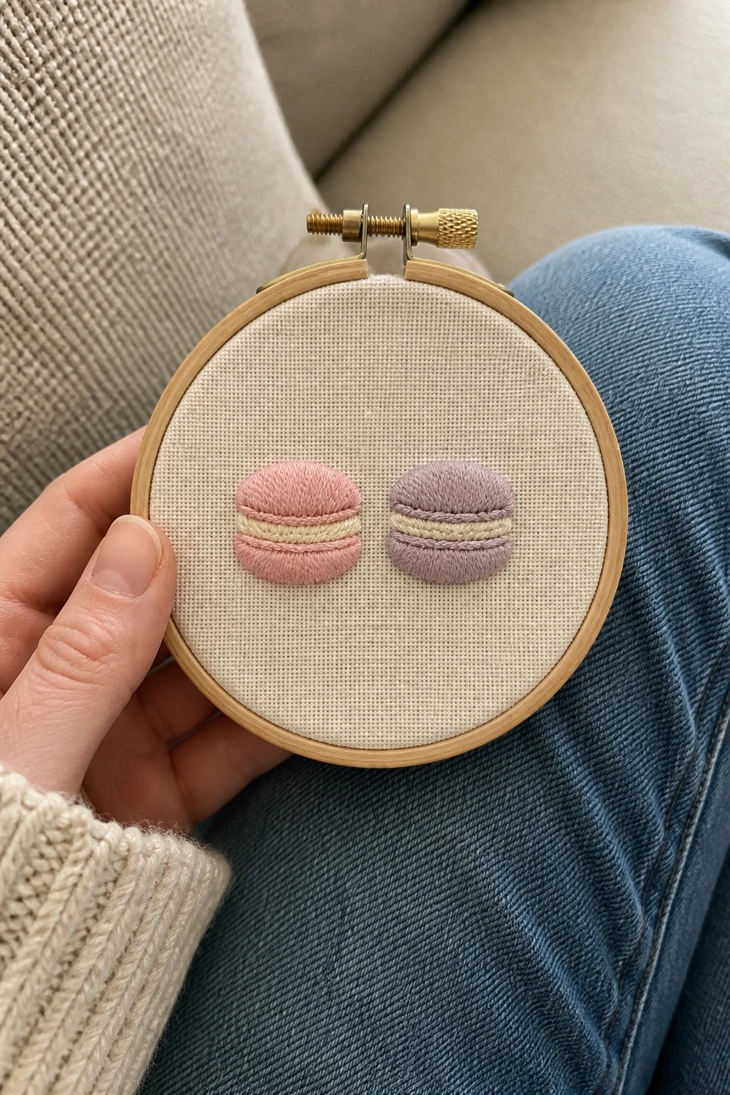Hand holding embroidery hoop with pink and lavender macarons over blue jeans