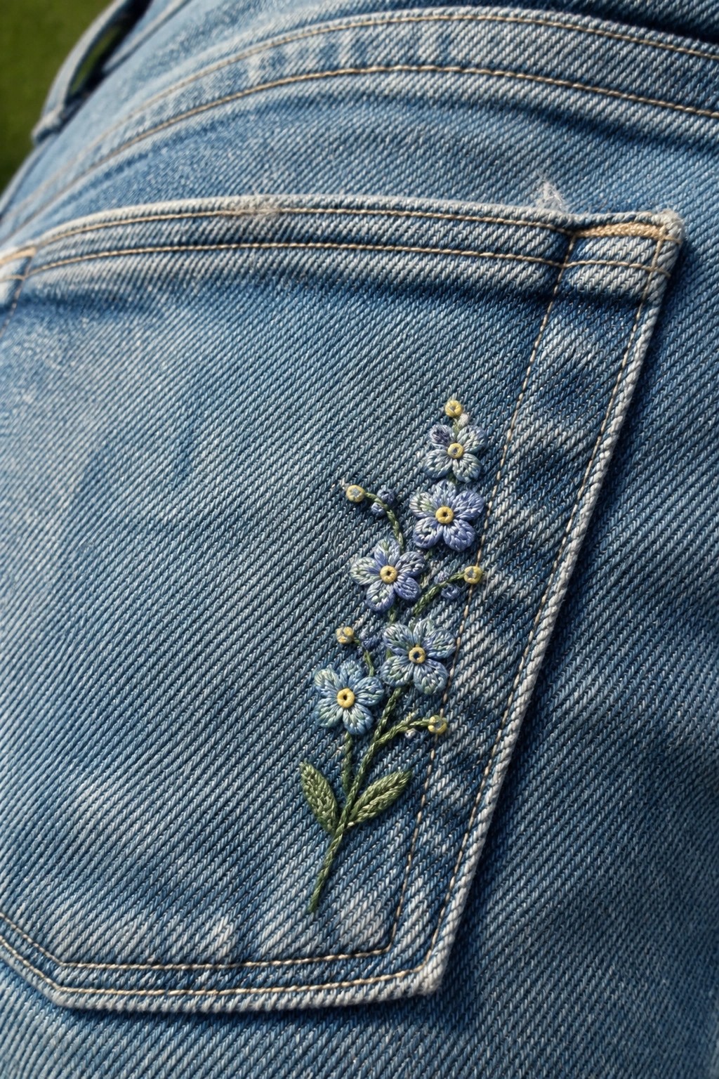 Blue denim jeans back pocket embroidered with small blue flowers and green stems.