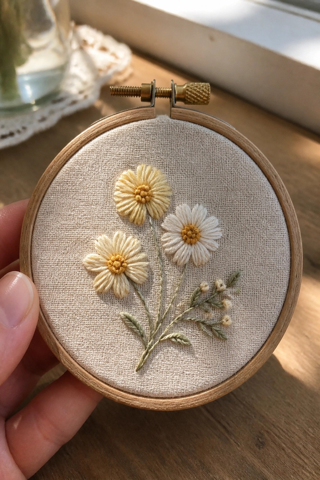 Hand holding embroidery hoop with three yellow and white daisies on beige fabric.