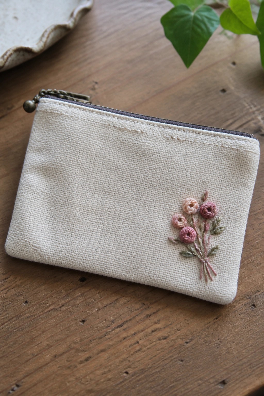 Cream-colored zip pouch with pink rose embroidery on rustic wooden table
