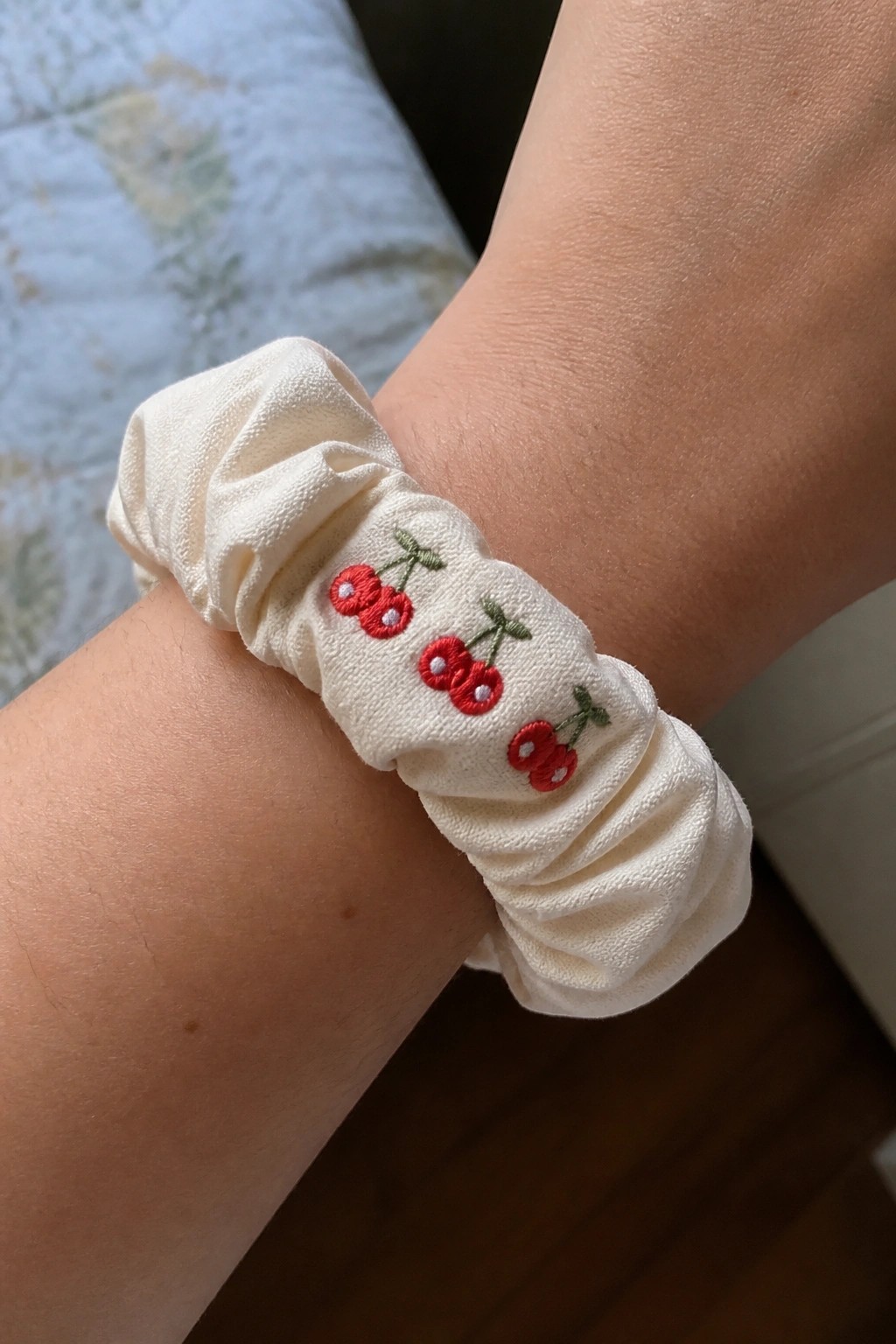 A cream fabric scrunchie with red embroidered cherries and green stems on a person's wrist.