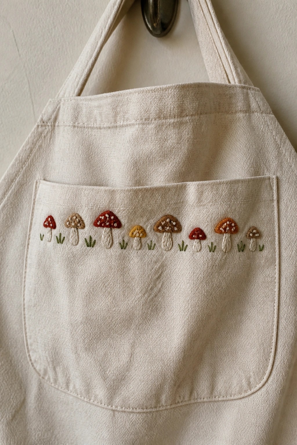 Cream apron pocket with row of colorful hand-embroidered mushrooms and grass.