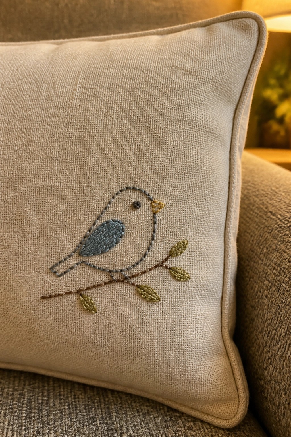 Beige pillow embroidered with blue bird on leafy brown branch with yellow beak.