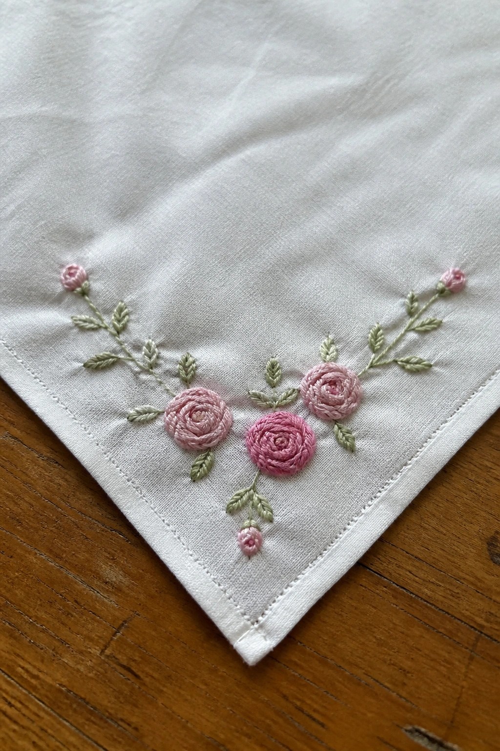 White cloth corner embroidered with pink roses, green leaves on wooden surface