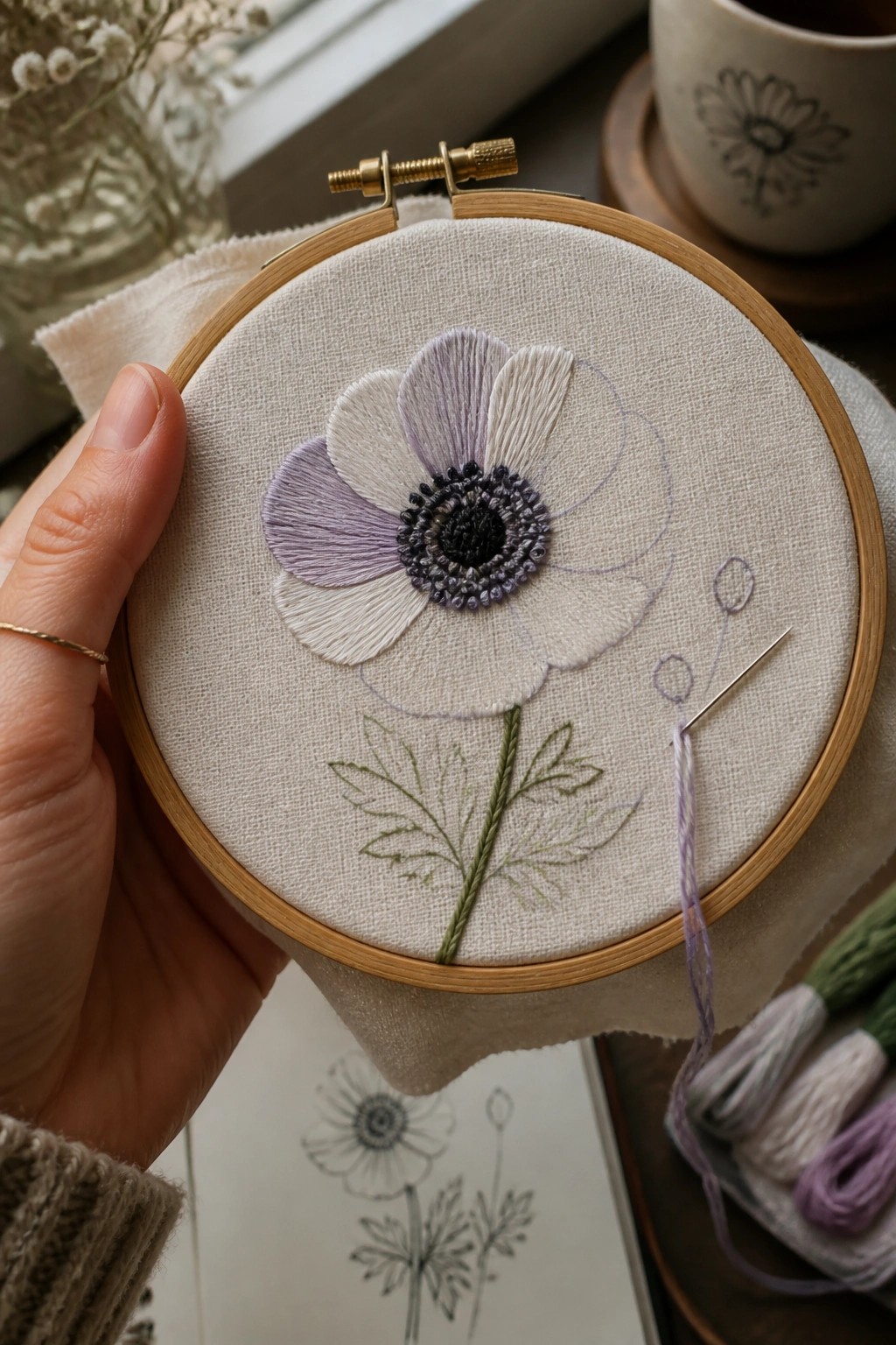 Hand holds hoop with purple-white flower embroidery, needle, and green stem.