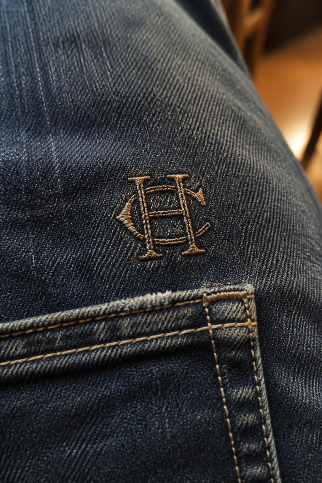 Blue denim jeans with brown embroidered monogram above stitched pocket edge.