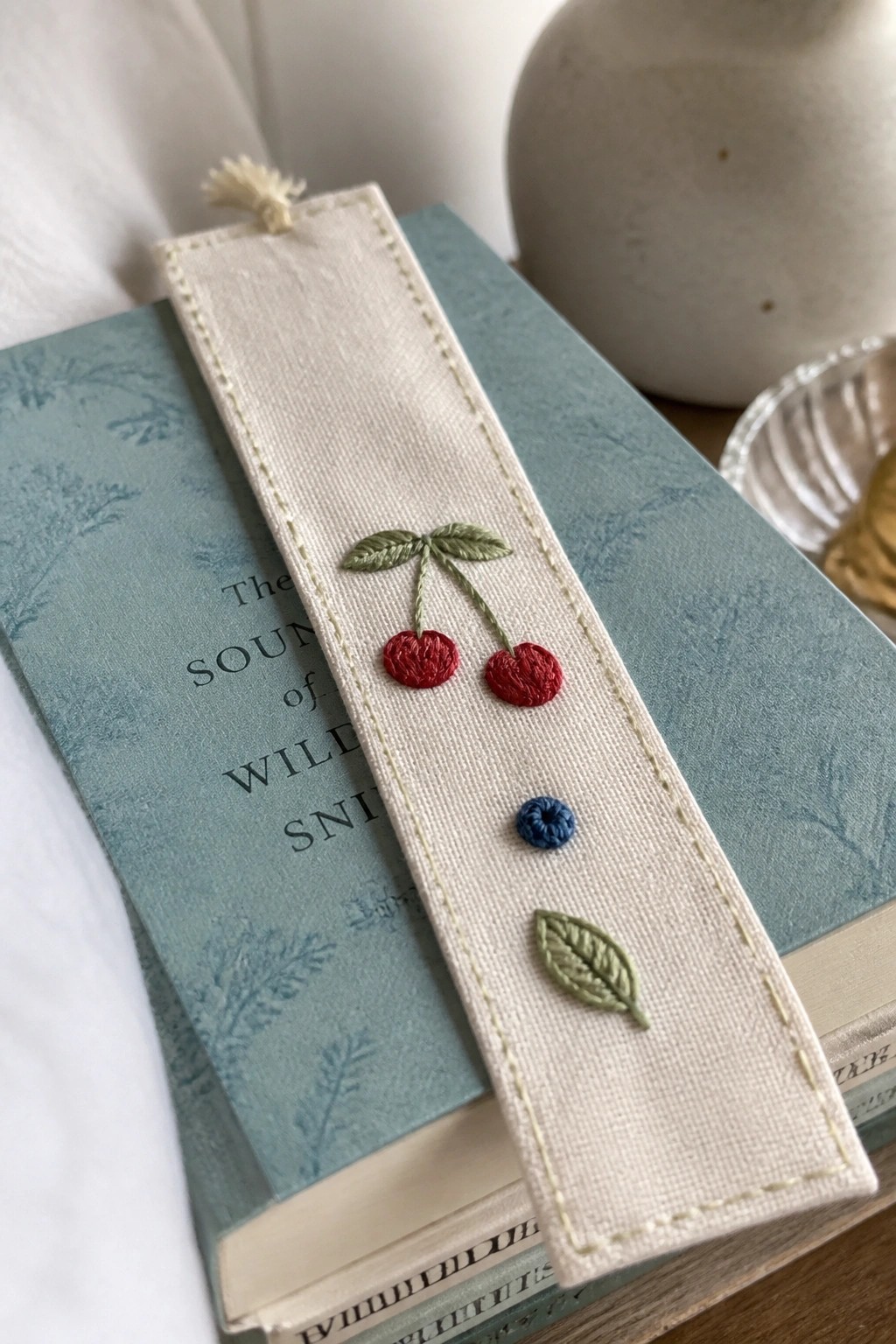 Embroidered cream bookmark with red cherries, green leaves, and blue dot on teal book