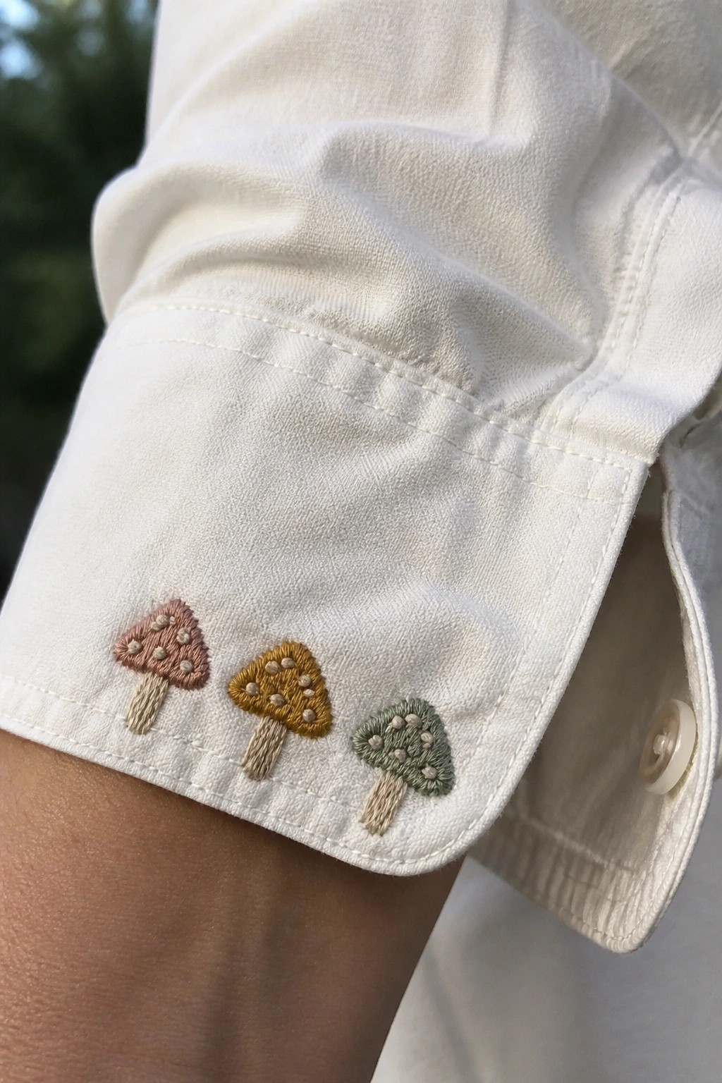 White shirt cuff with three embroidered mushrooms in pink, yellow, and green.