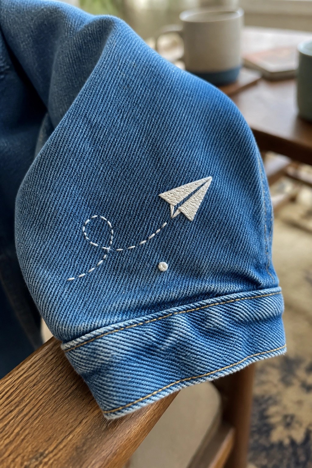 Blue denim jacket sleeve with embroidered paper airplane, dashed path, and button detail