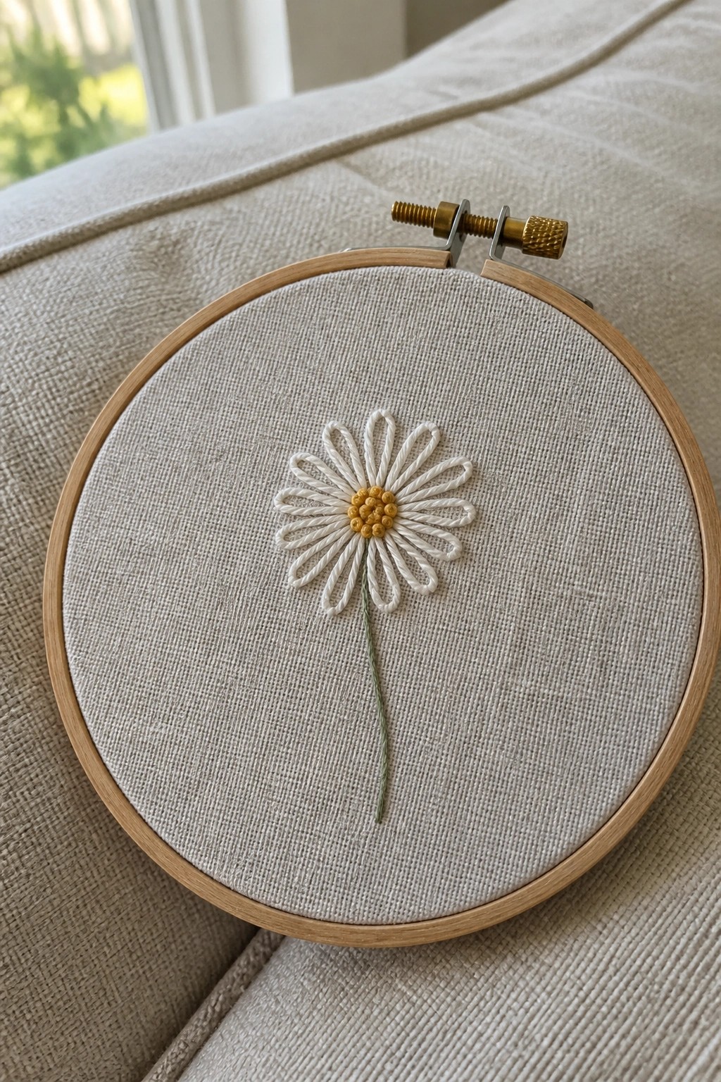 Wooden embroidery hoop with white daisy, yellow center, and green stem on beige fabric.