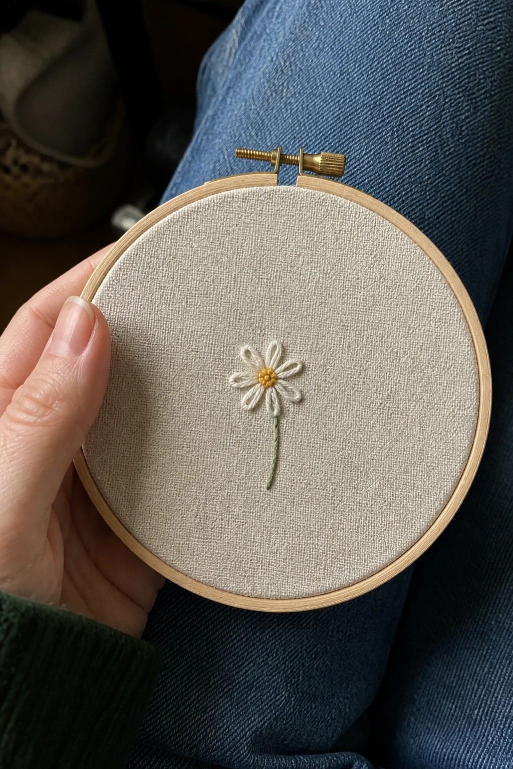 Hand holding wooden hoop with embroidered white daisy over blue jeans and green sleeve