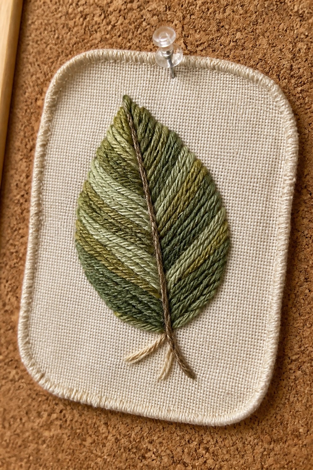 Green embroidered leaf on white fabric pinned to brown corkboard with pushpin.