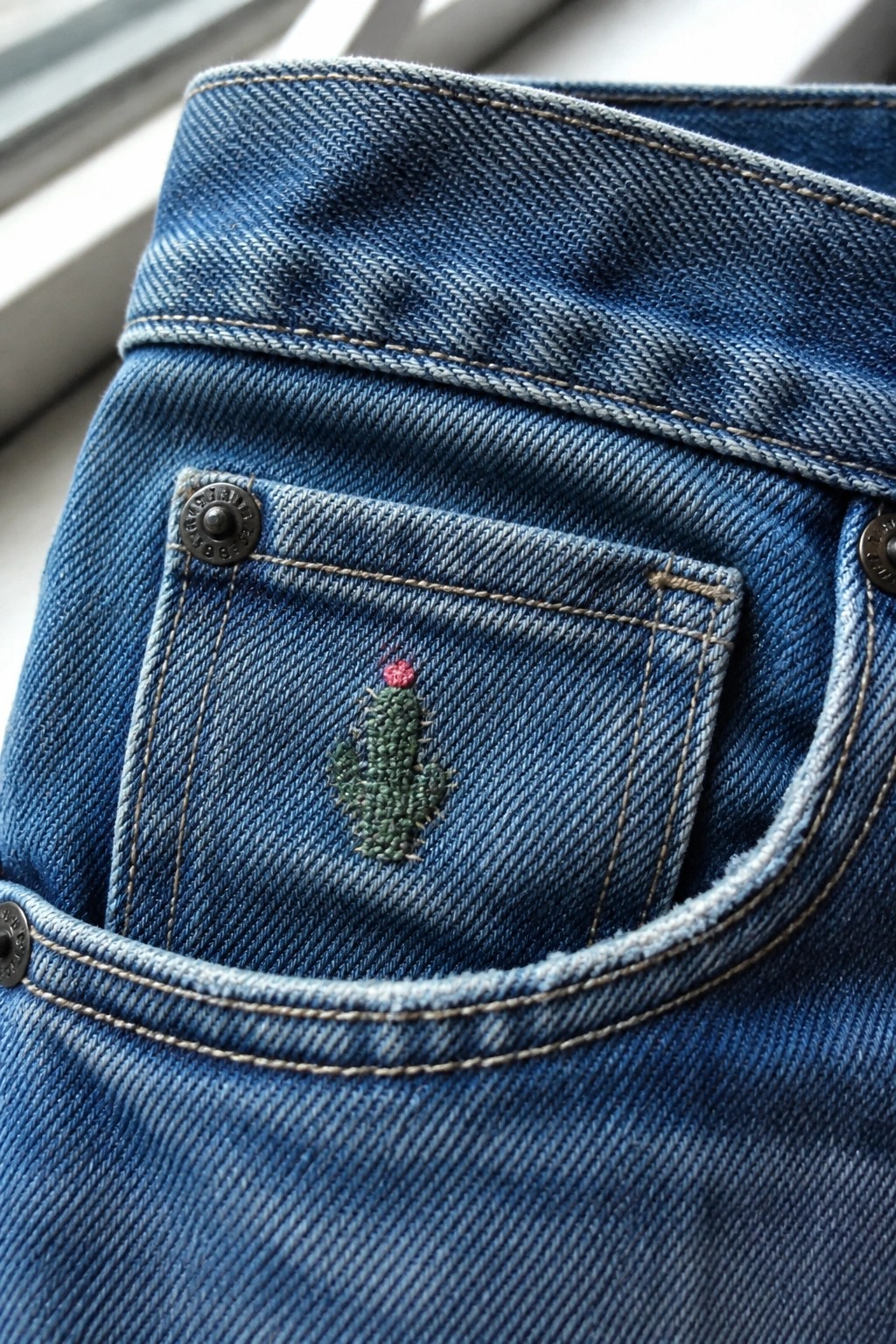 Blue denim jeans pocket with green embroidered cactus and metal rivets.