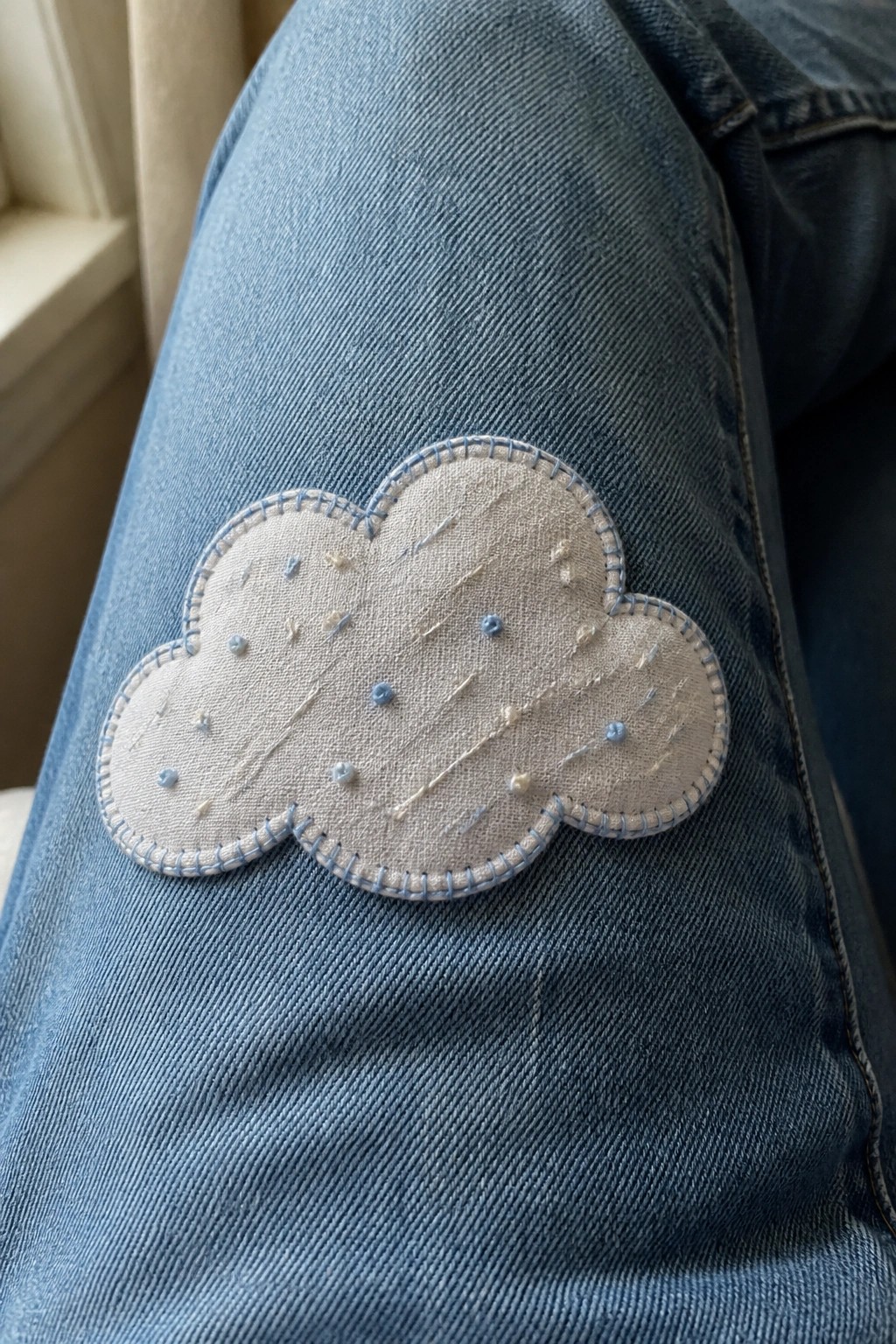 A light beige cloud-shaped fabric patch with blue stitching is sewn onto blue denim jeans.