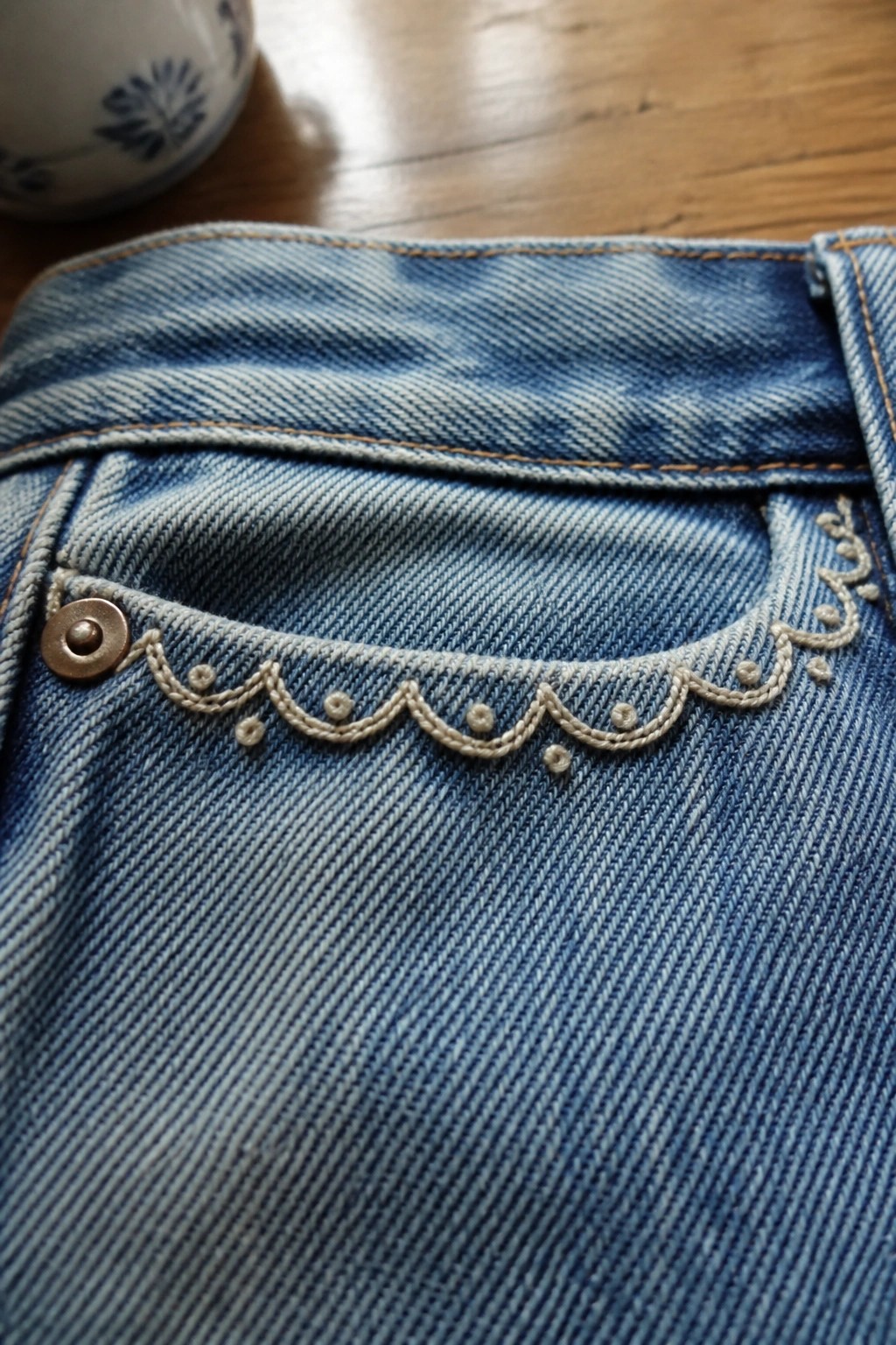 Blue denim jeans pocket with scalloped beige embroidery and metal rivet on wood