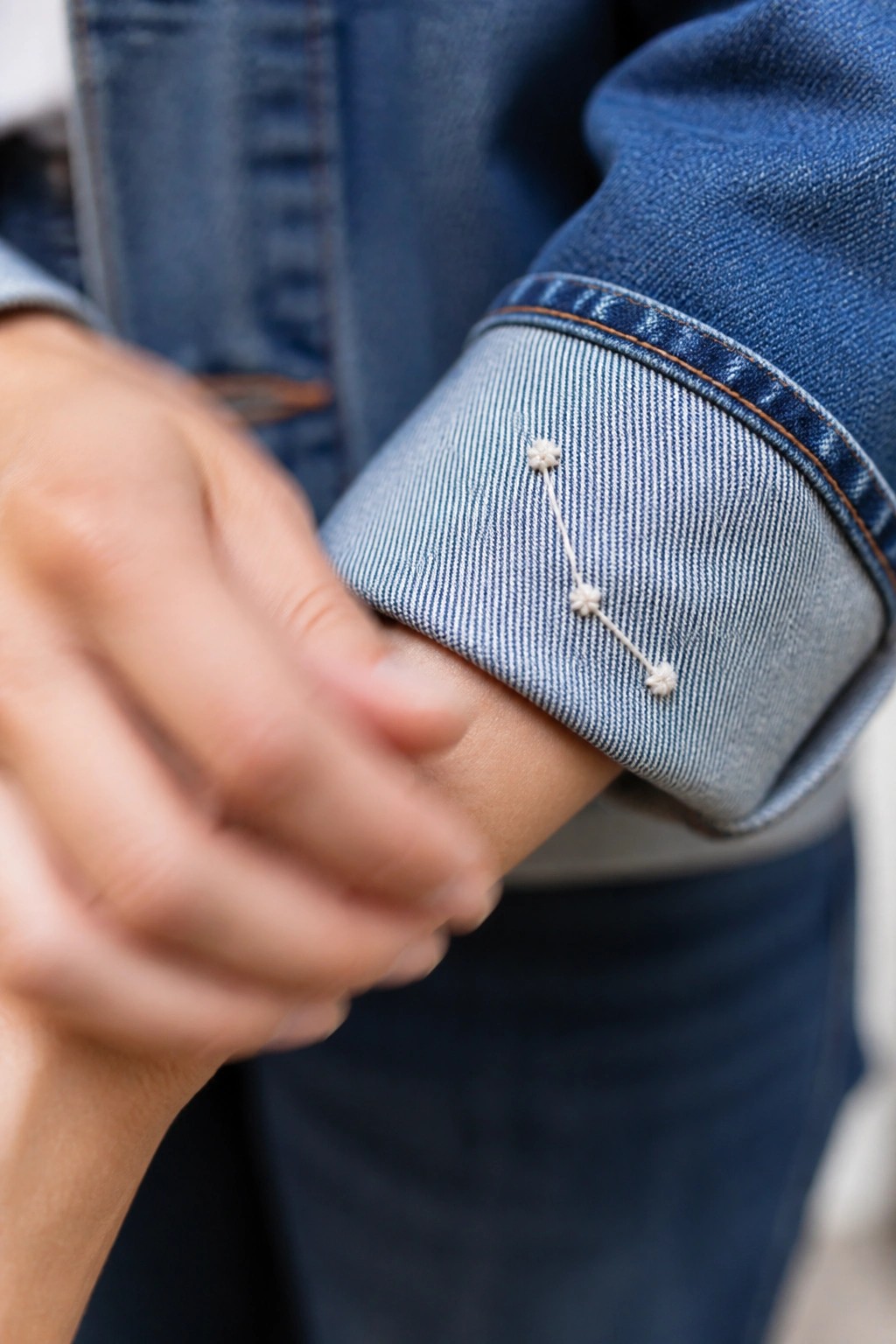 Blue denim jacket cuff with white embroidered stars connected by threads near a hand.
