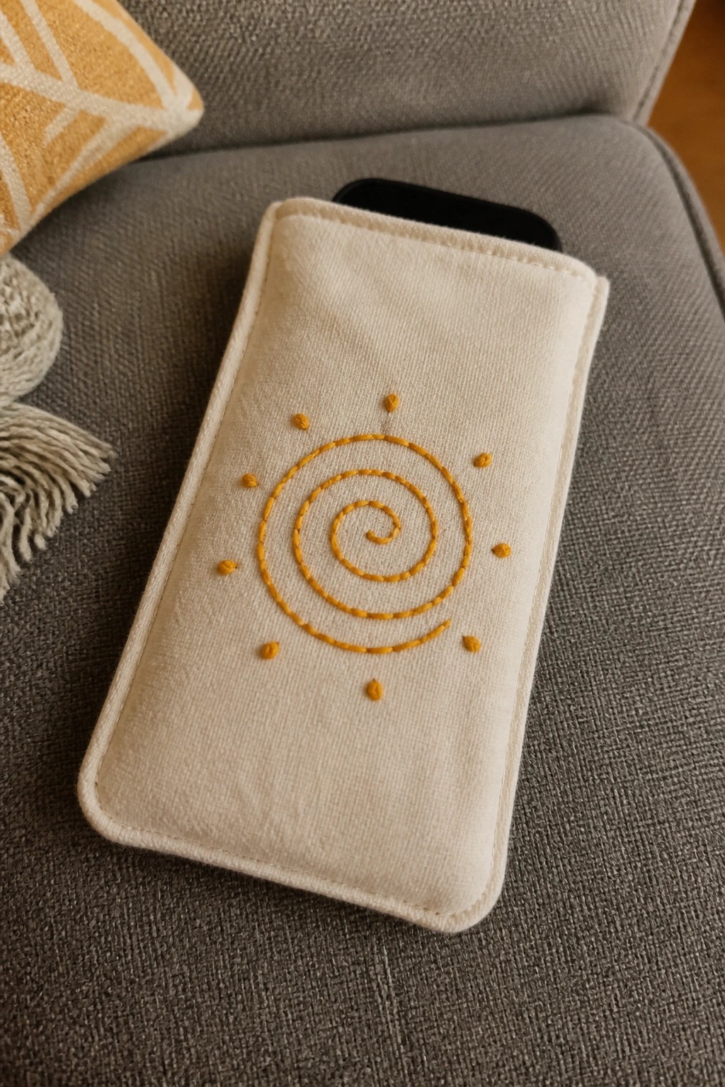 Cream fabric phone sleeve with yellow spiral sun embroidery on gray upholstery