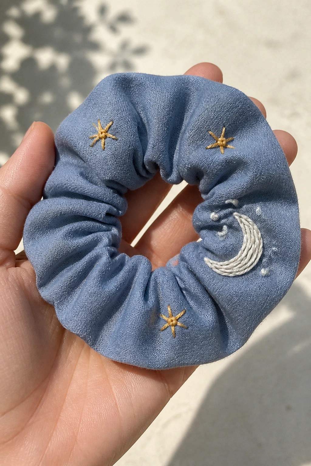 Hand holding light blue scrunchie embroidered with gold stars and white moon