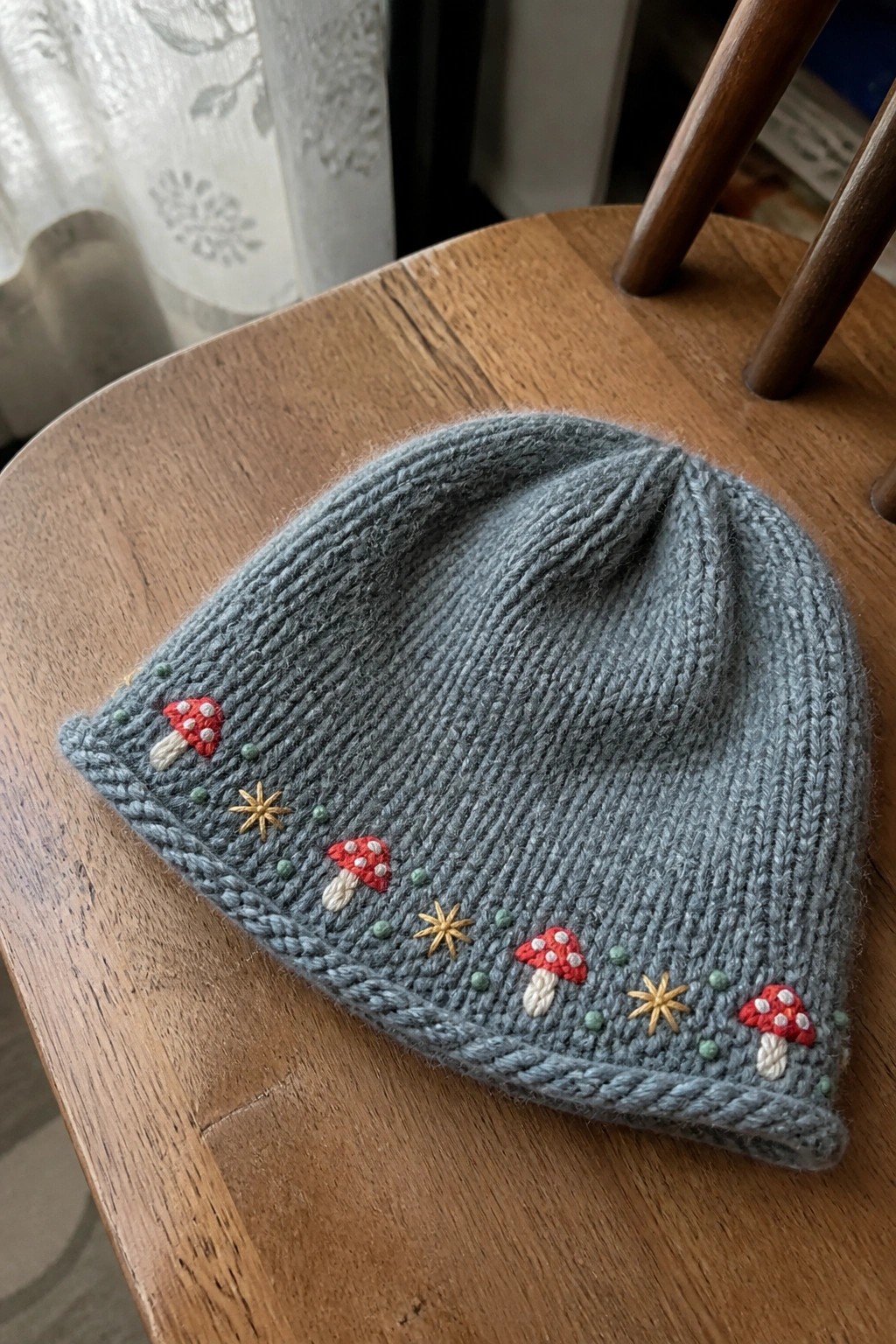 A gray knitted beanie embroidered with red mushrooms, gold stars, and green dots resting on a wooden chair.