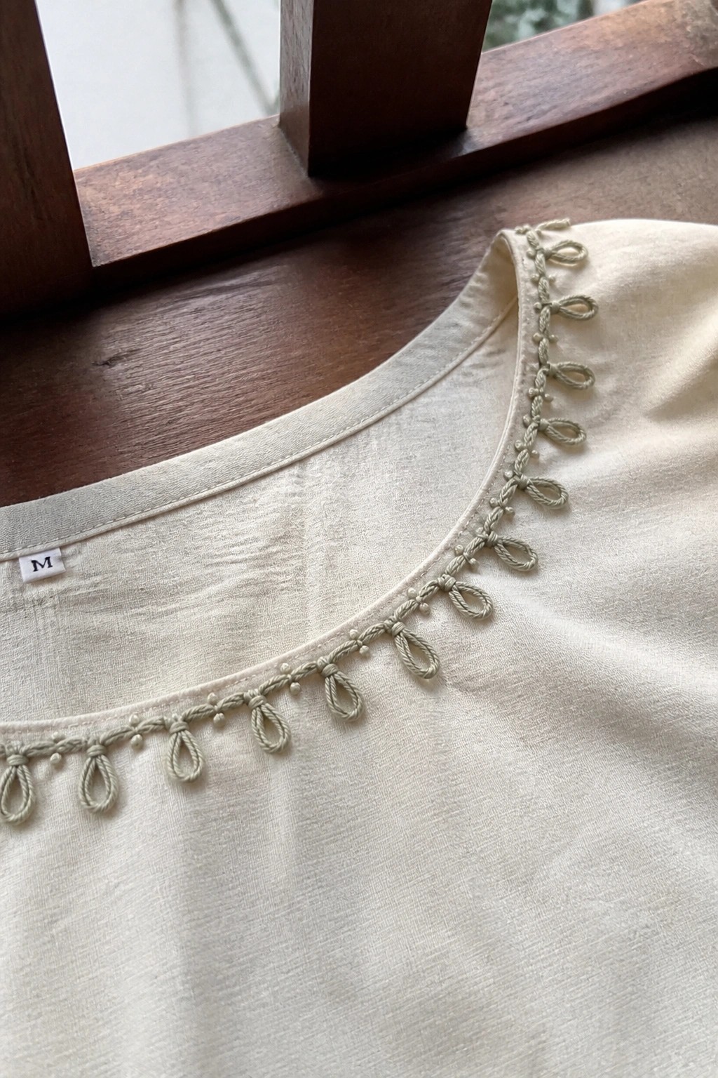 Close-up of cream fabric neckline with light green looped embroidery along the curved edge.
