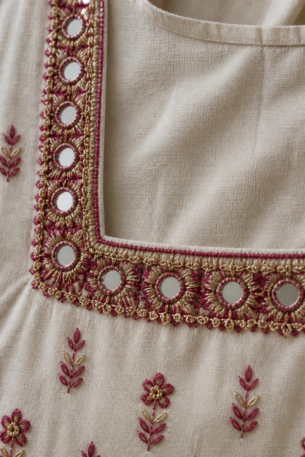 Close-up of beige fabric with a square neckline edged in gold and magenta embroidered circles holding small mirrors, plus scattered floral motifs below.