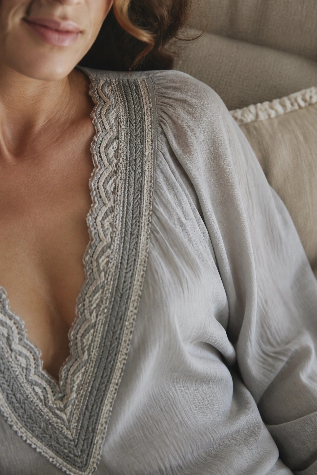Woman in light gray blouse with intricate embroidery on V-neck, reclining on beige couch