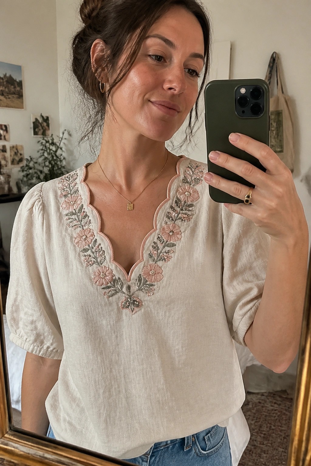 Woman in cream blouse with pink floral embroidery taking mirror selfie with green phone.