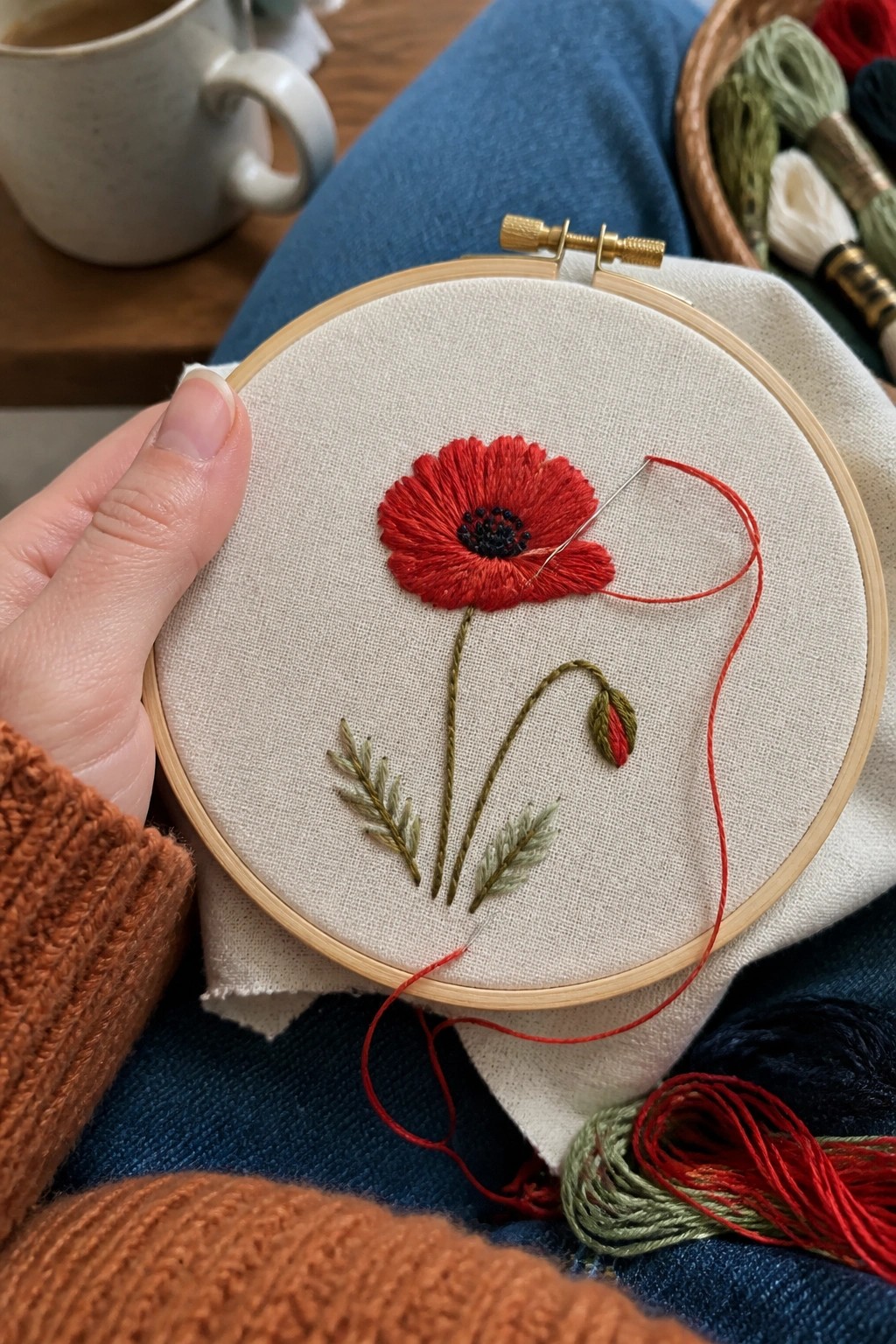 Hand in orange sweater holding hoop with red poppy, needle, and thread on fabric