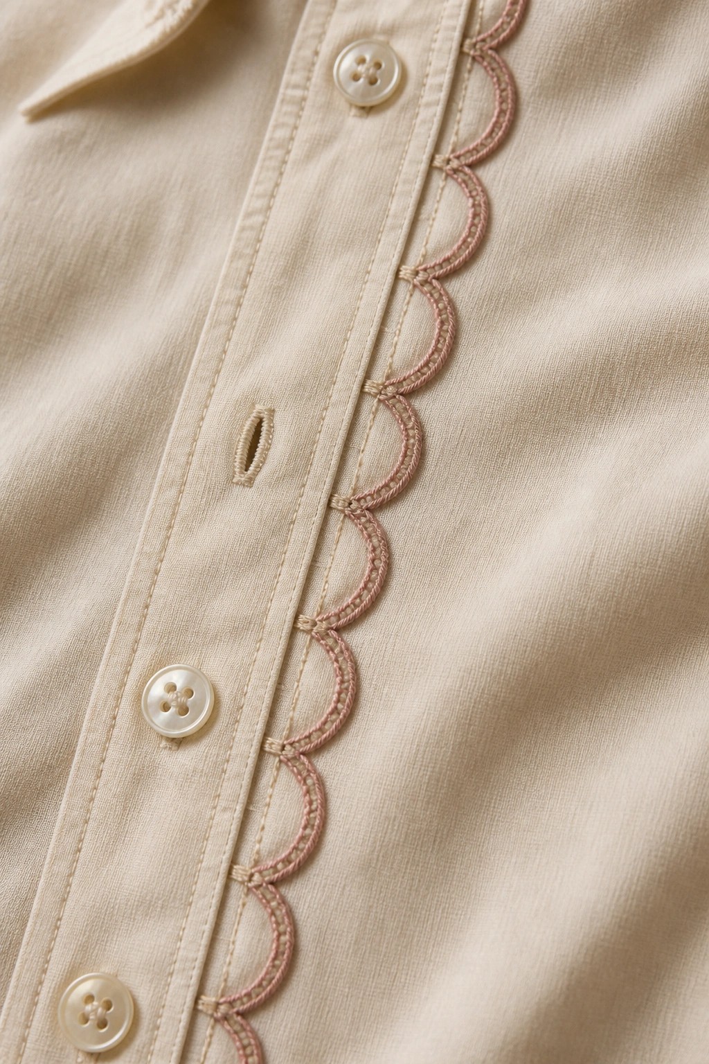 Close-up of a beige shirt with pink scalloped embroidery running beside the button placket and white buttons.