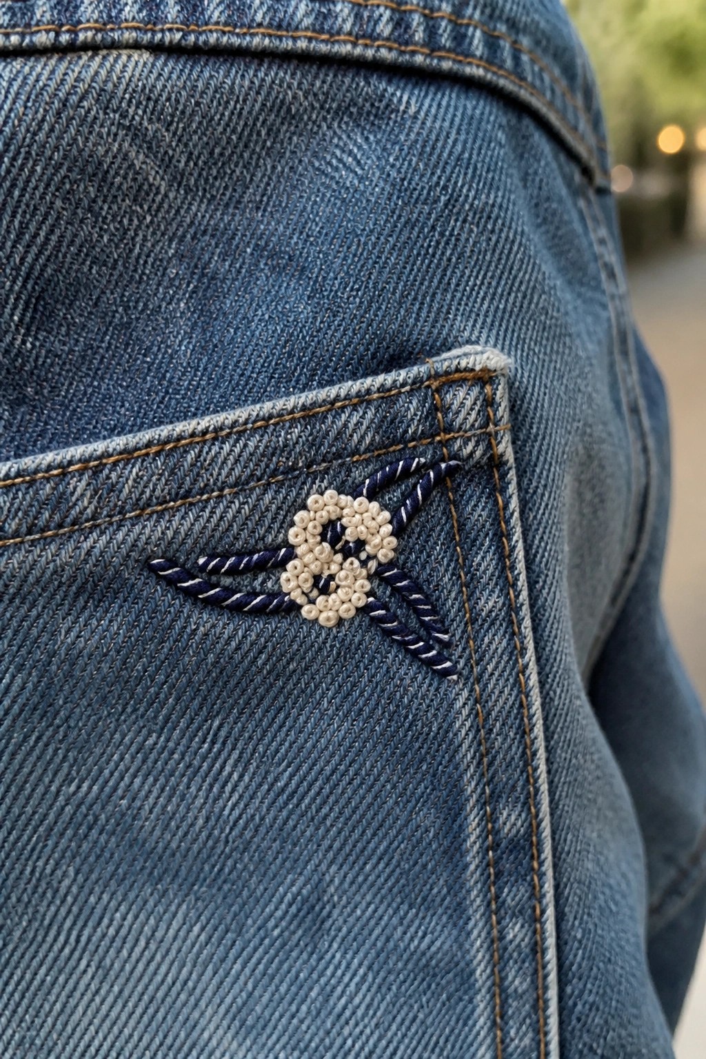 Blue denim jeans pocket with white beaded flower and blue thread embroidery.