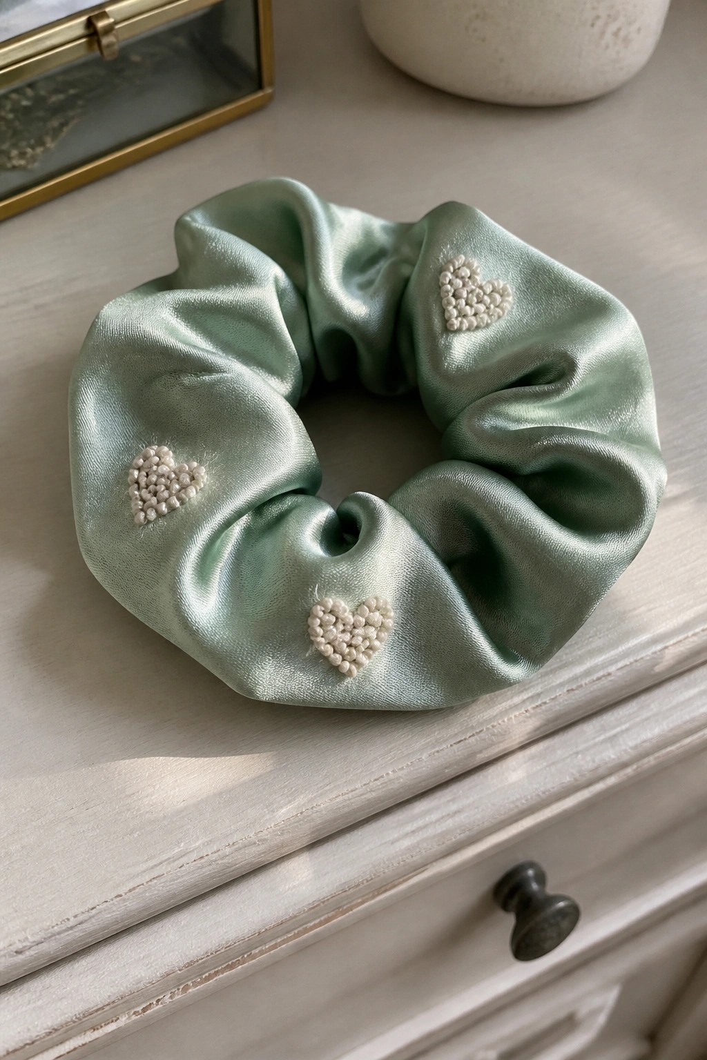 A mint green satin scrunchie with three small white hearts formed from clustered knots embroidered on the fabric.
