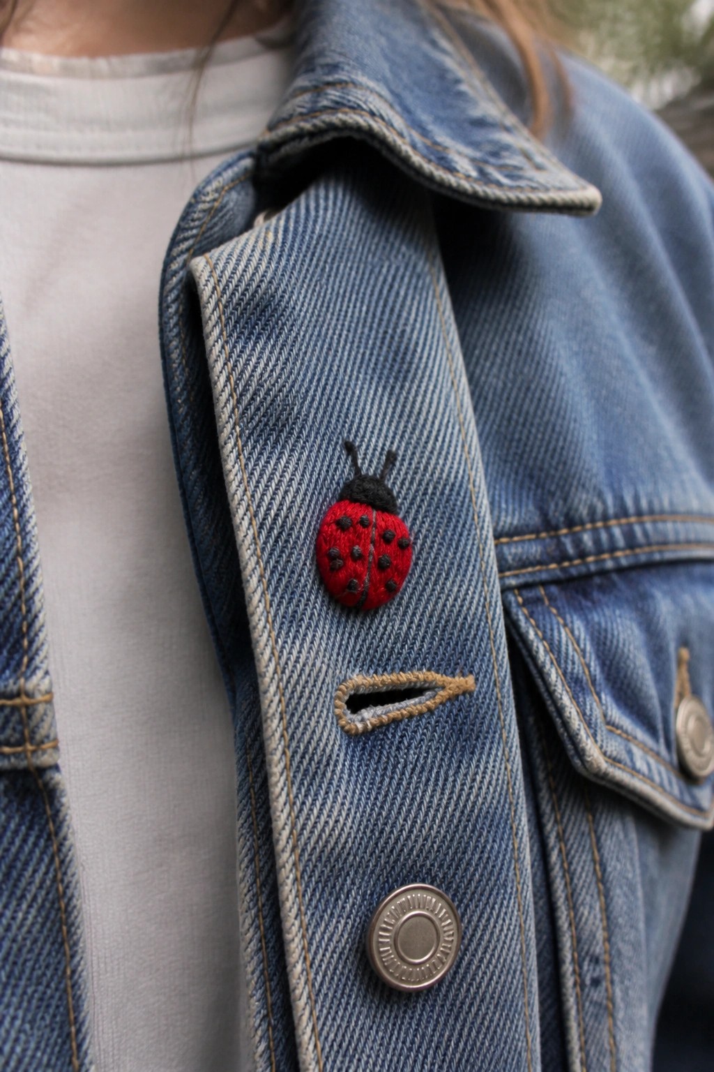 Red embroidered ladybug on blue denim jacket lapel above metal button.