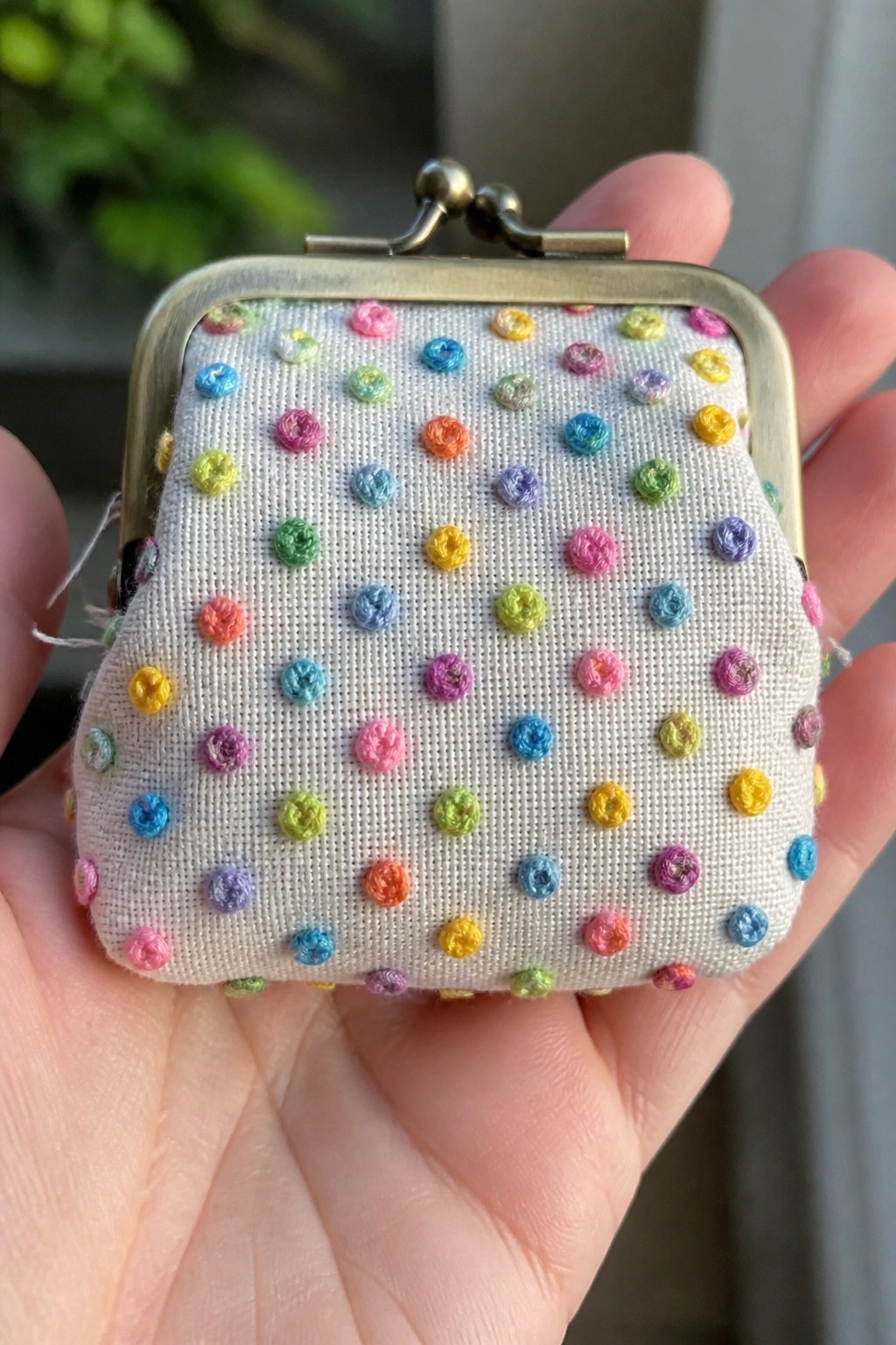 Hand holding white fabric coin purse with colorful embroidered dots and metal clasp.