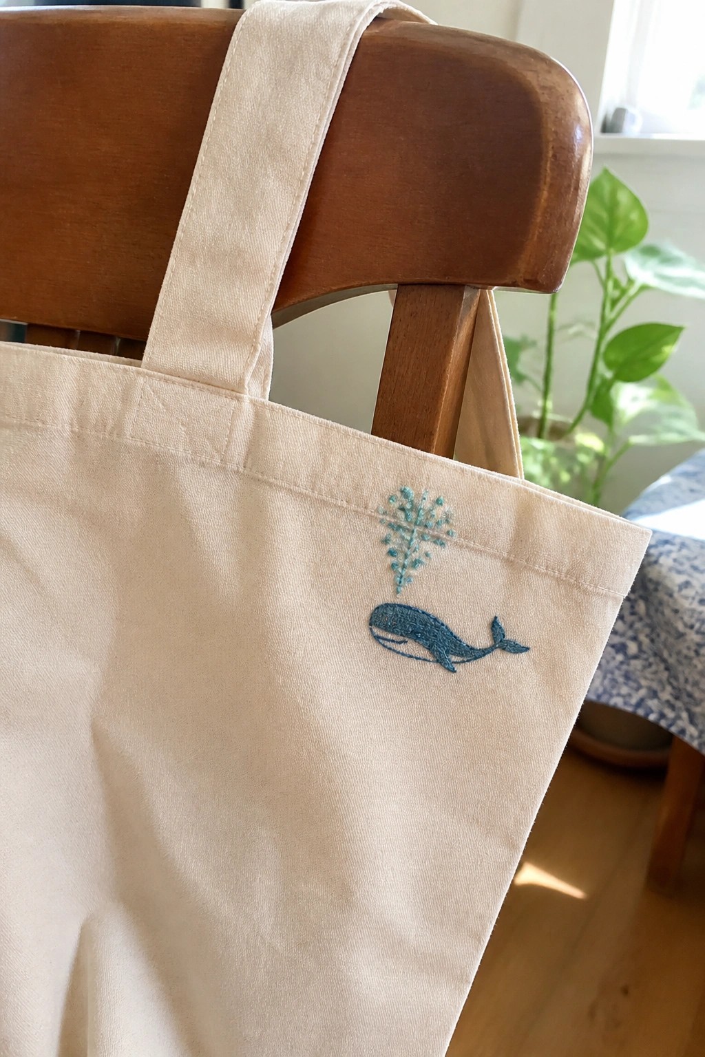 Cream canvas tote bag with blue whale embroidery draped over wooden chair.