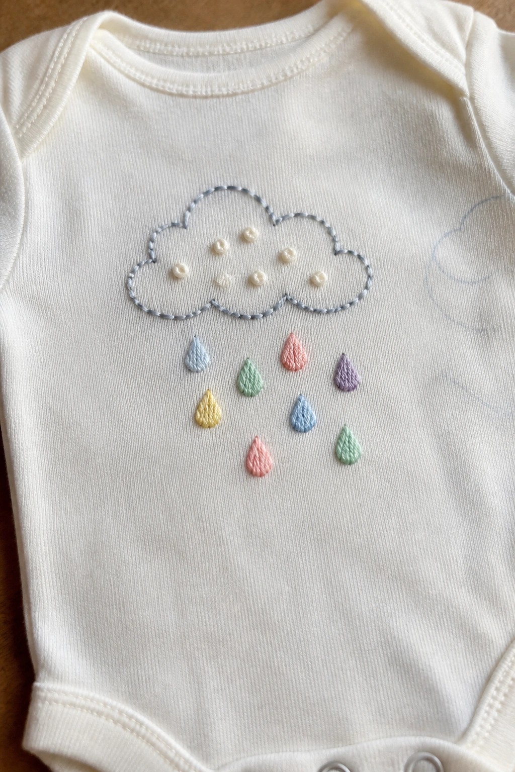 White baby onesie with embroidered cloud, buttons, and pastel rainbow raindrops.