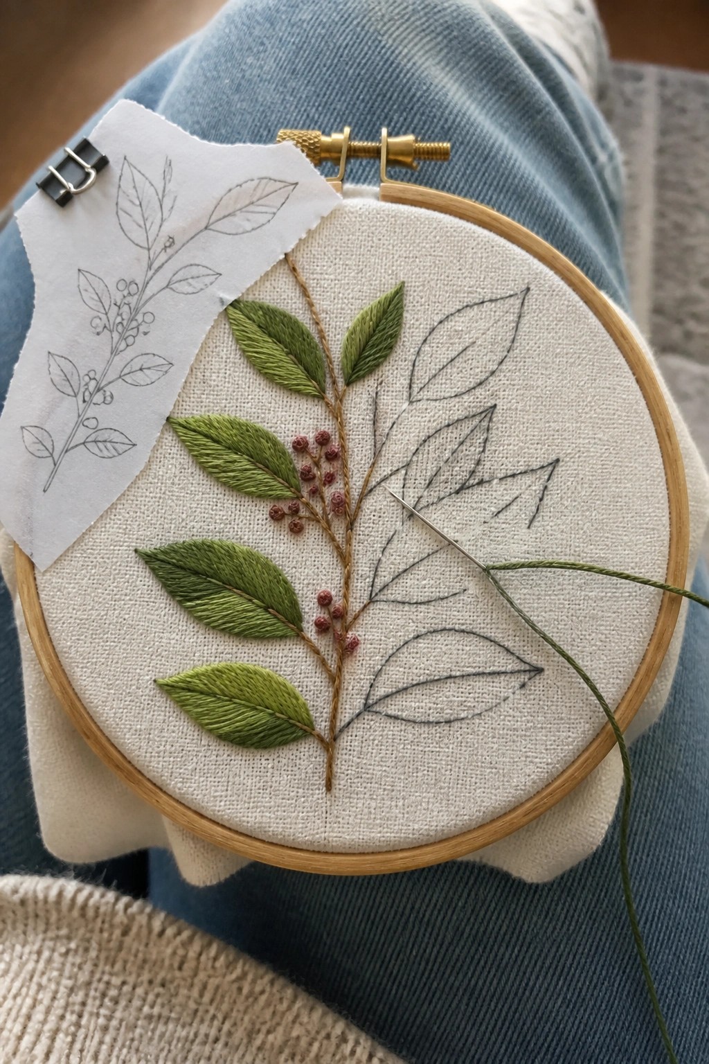 Embroidery hoop on blue jeans with green leaves, red berries, needle, and paper pattern