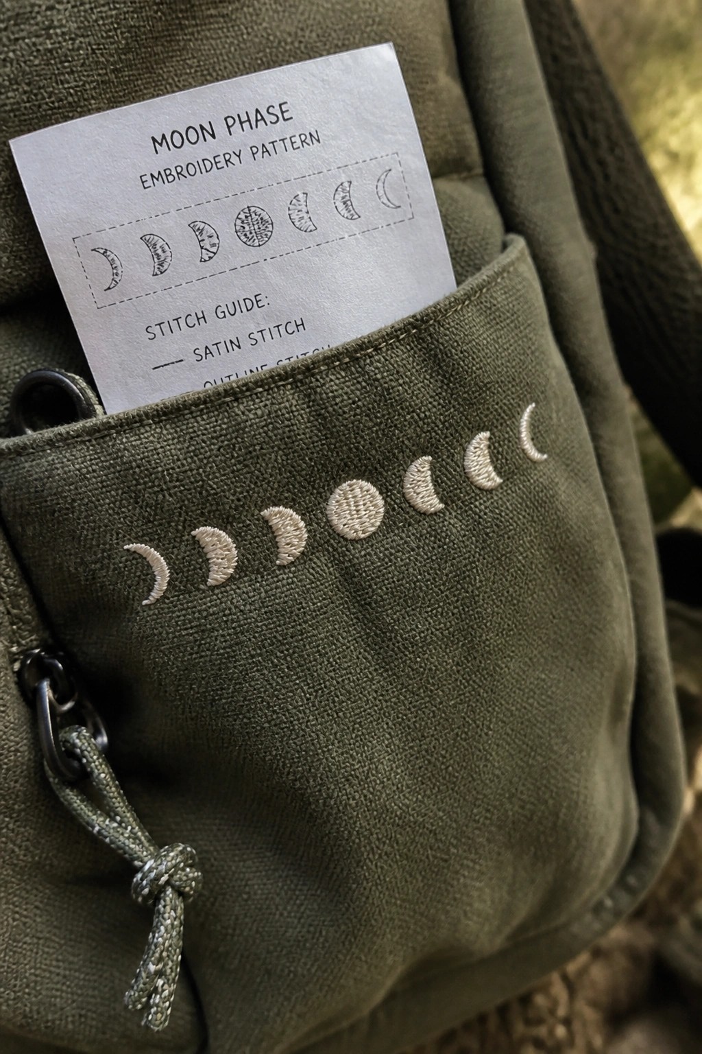 A green canvas bag pocket with white moon phase embroidery and a paper pattern card tucked inside.