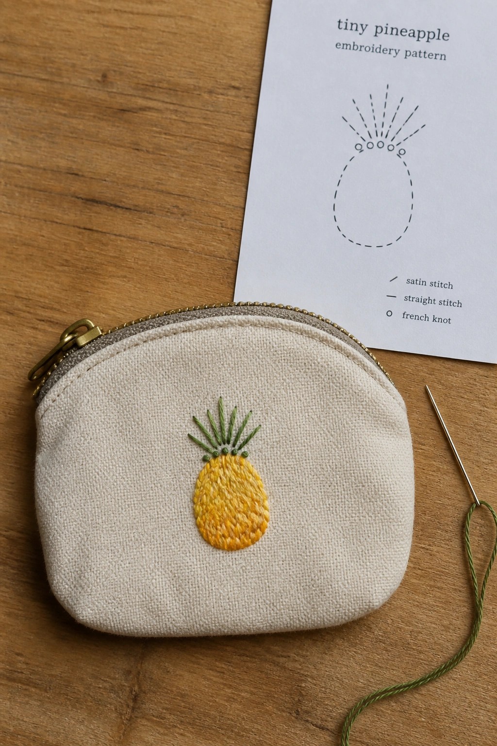 Cream zippered pouch with yellow pineapple embroidery beside pattern sheet and needle on wood