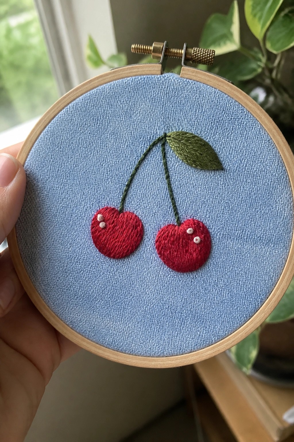 Hand holds wooden hoop with two red cherries embroidered on blue fabric