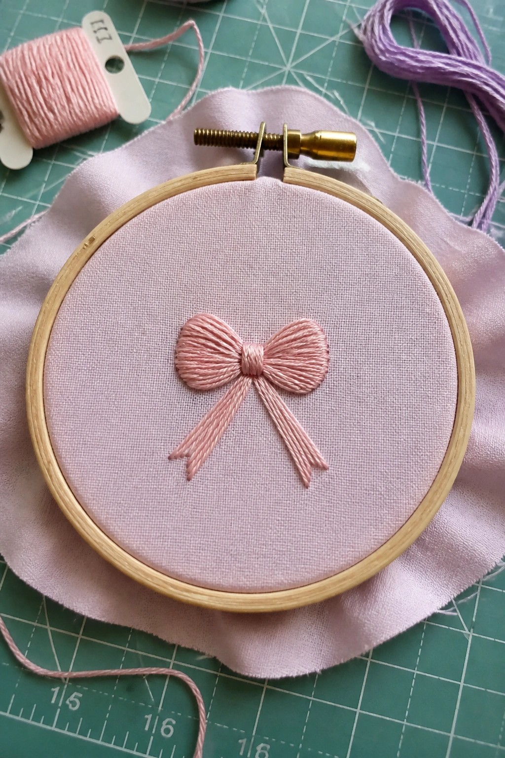 Pink embroidered bow on fabric in wooden hoop on green cutting mat