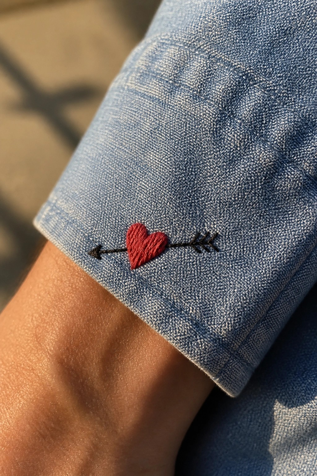 Blue denim shirt cuff with red heart pierced by black arrow embroidery.