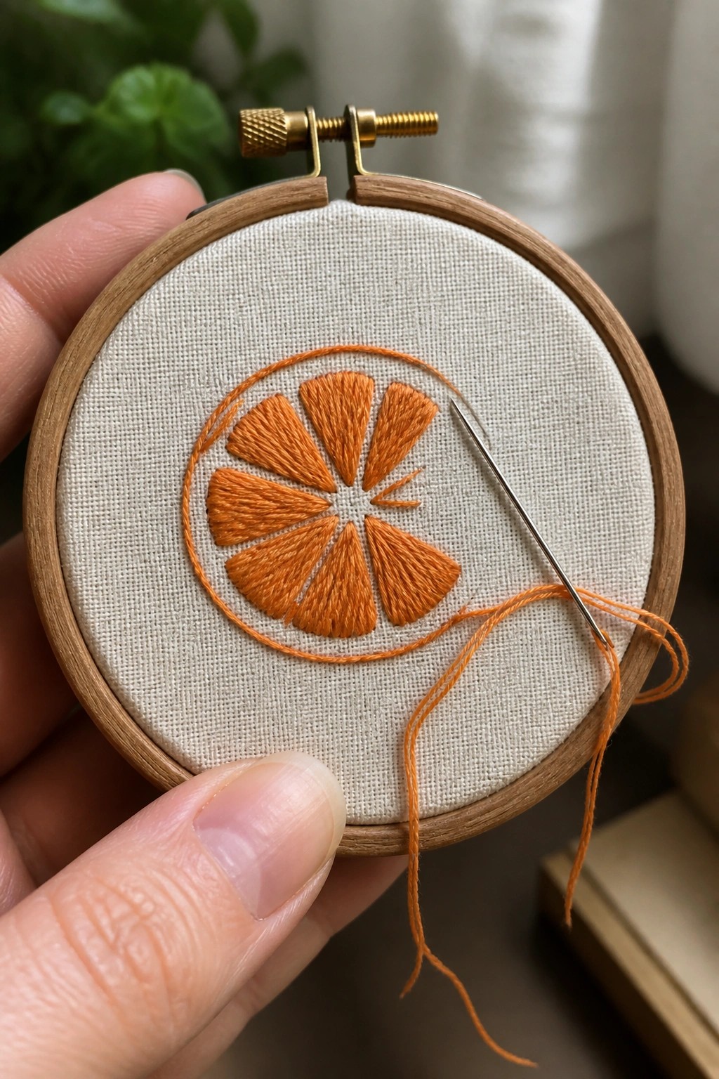 Hand holding wooden hoop with orange citrus slice embroidery and dangling needle.