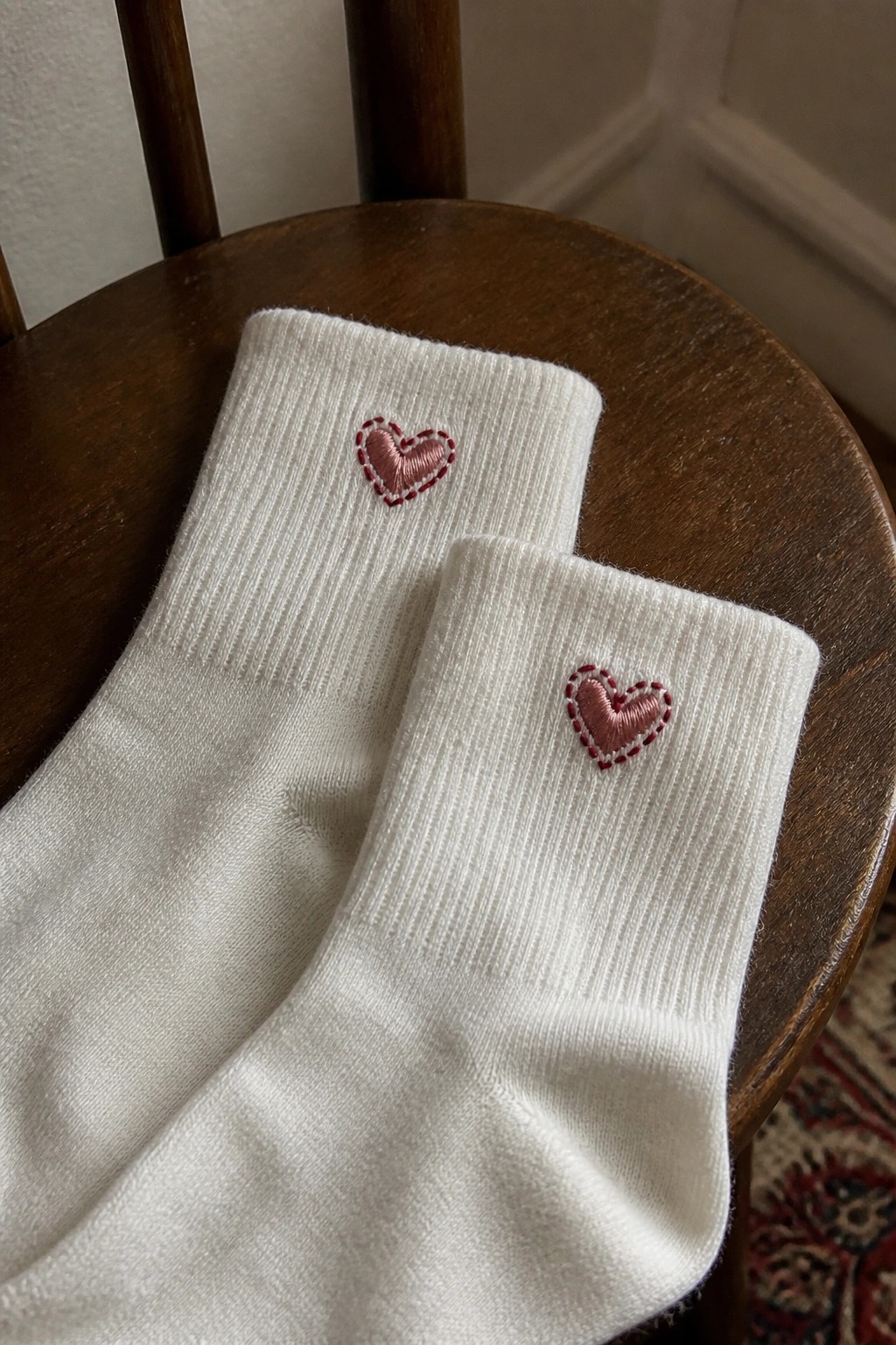 Two white socks with small pink hearts embroidered on the ankles resting on a wooden chair.