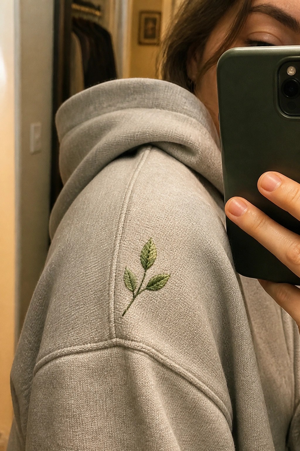 Gray hoodie with green embroidered leaves on shoulder, hand holding smartphone.