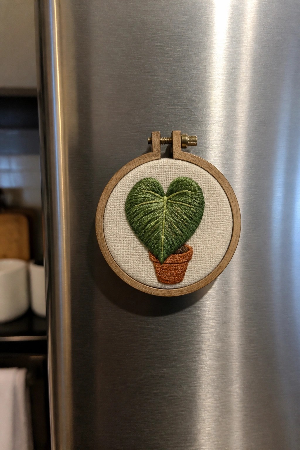 Embroidered heart-shaped green leaf in terracotta pot within wooden hoop on refrigerator