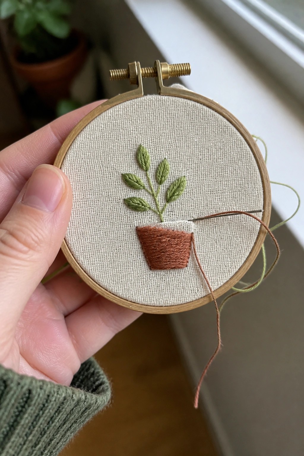 Hand in green knit sleeve holds hoop with embroidered potted plant and needle