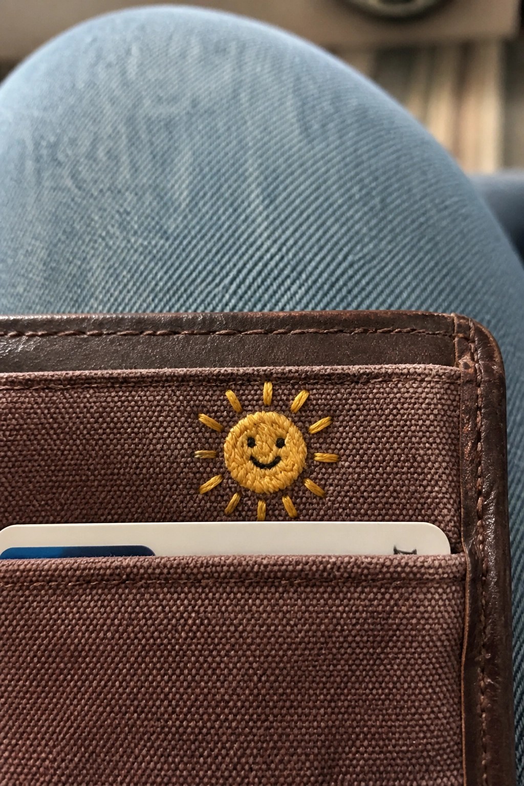 Brown wallet with yellow embroidered smiling sun and white card on blue denim.