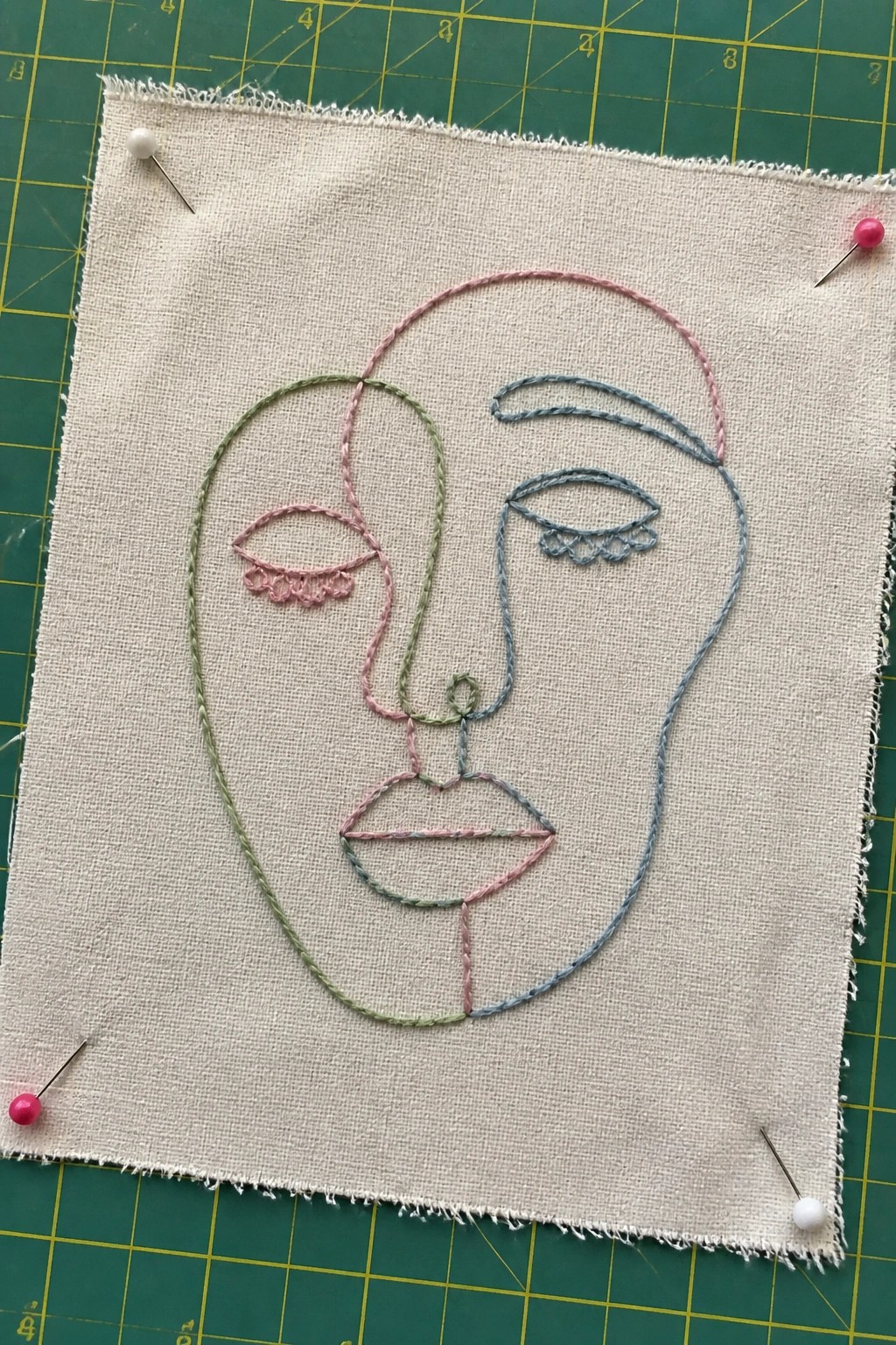 Embroidered abstract face in pink, green, and blue threads on beige fabric